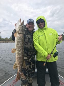 Two men on a boat smile while one holds up a large fish and the other holds a fishing rod, with water in the background.