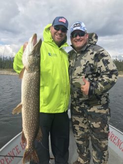 Two men on a boat pose with a large fish catch; both are wearing hats and sunglasses, with water and trees in the background.