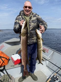 A man in a boat holds up two large fish, smiling, with water and fishing gear visible in the background.