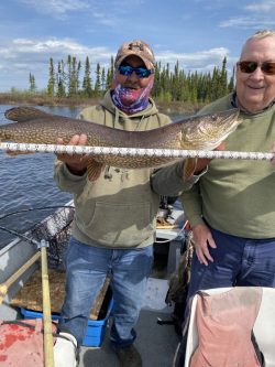Two men in a boat hold a large fish against a measuring stick on a lake with trees in the background.