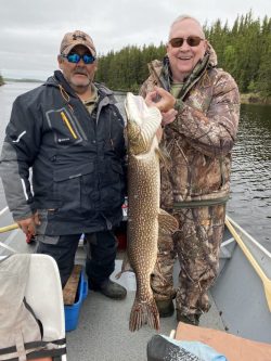 Two men in outdoor gear stand on a boat holding a large fish they caught, with trees and water in the background.