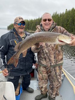 Two men in outdoor gear stand in a boat, one holding a large fish with a forest and lake in the background.