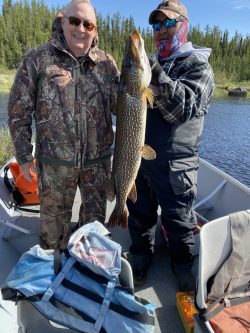 Two people in outdoor gear on a boat display a large fish they caught, with trees and water in the background.