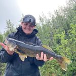 Person in a black jacket and cap holding a large fish outdoors with trees in the background.