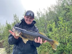 Person in a black jacket and cap holding a large fish outdoors with trees in the background.