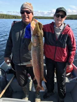 Two people standing on a boat hold up a large fish, with a lake and trees visible in the background.