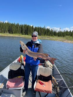 Person standing in a boat on a lake, holding a large fish with trees and sky in the background.