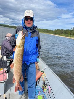 Person in blue jacket holding a large fish on a boat in a lake, with another person sitting in the background.
