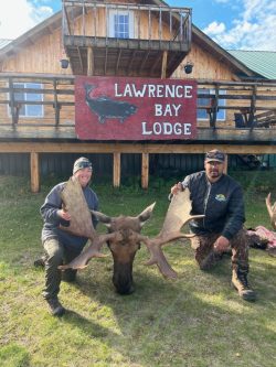 Two people kneel beside a large moose antler rack in front of a lodge with a "Lawrence Bay Lodge" sign.