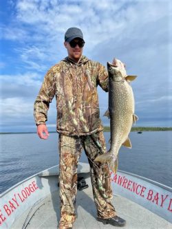 Person in camouflage clothing stands on a boat holding a large fish, with water and sky in the background.