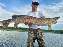 Man in sunglasses and camo pants holding a large northern pike fish near a lake with trees in the background.