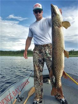 Man standing on a boat holding a large fish beside him, with a lake and trees in the background.