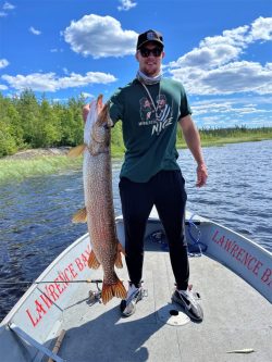 Man standing on a boat holding a large fish with a lake and trees in the background.