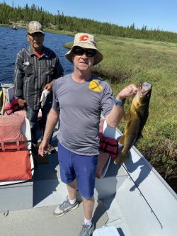 Two men stand on a boat; one holds a large fish beside a river with grassy banks and trees in the background.