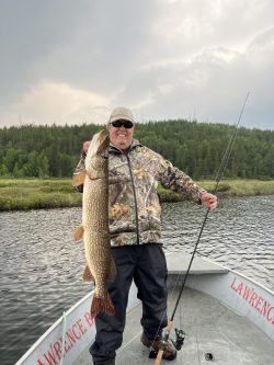 Person standing on a boat holding a large fish with one hand and a fishing rod with the other, surrounded by water and trees.