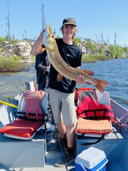 A person standing on a boat holds a large fish with one hand, smiling at the camera on a sunny day.