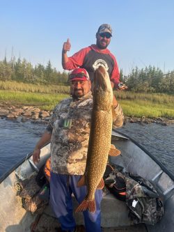 Two men on a boat hold up a large fish they caught, with one man giving a thumbs-up. Trees and shoreline are in the background.