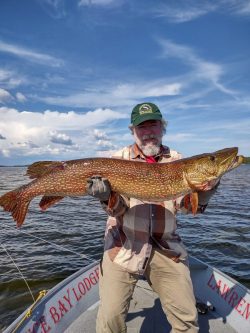 A man standing on a boat holds a large fish with both hands under a partly cloudy sky.