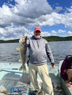 Person standing on a boat holding a large fish, with a lake and forested shoreline in the background under a cloudy sky.