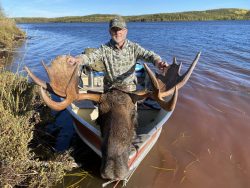 A man in camouflage holds a large moose head with antlers in a boat on a lake, with forested hills in the background.