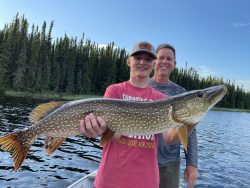 Two people on a boat hold a large fish in front of a forested lake under a blue sky.