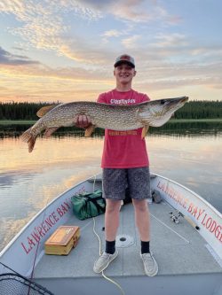 Young man standing on a fishing boat holding a large fish with both hands, with a lake and forest in the background.
