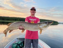 A person on a boat holds a large northern pike fish with a lake and treeline in the background at sunset.
