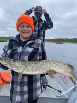 A boy in a plaid jacket and orange hat holds a large fish on a boat, with another person standing behind him.