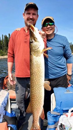 Two smiling men on a boat holding a large fish, with trees and water in the background.