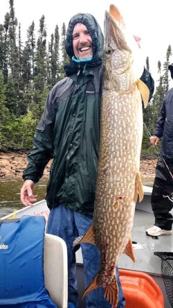 A man in a raincoat holds a very large fish on a boat, with trees and water visible in the background.