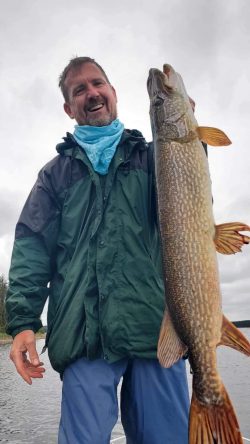 A man in outdoor clothing holds up a large fish by the water on a cloudy day.
