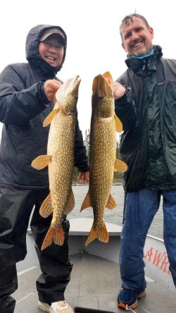 Two people in rain gear holding large northern pike fish on a boat, smiling at the camera.