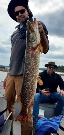 A man standing in a boat holds up a large fish, while another man sits behind him looking at his phone.