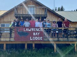 A group of people stand on a wooden deck in front of a lodge with a sign that reads "Lawrence Bay Lodge.