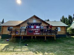 A group of people stand on the deck of a wooden building with a sign reading "Lawrence Bay Lodge.