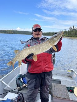 A person in a red jacket holds a large fish on a boat, with a lake and forest in the background.