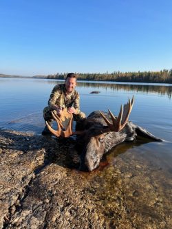 A man in camouflage kneels beside a large moose lying on the shore of a lake with a forest in the background.