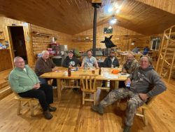 Seven men sit around a wooden table in a log cabin, with food, drinks, and papers visible on the table.