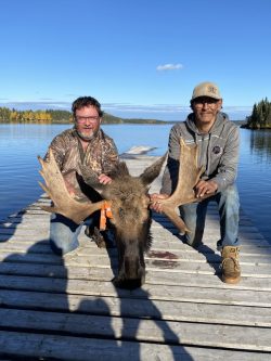 Two men kneel on a dock by a lake, holding the head and antlers of a harvested moose.