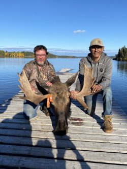 Two men kneel on a dock by a lake, holding a moose head with large antlers between them, trees visible in the background.