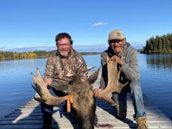 Two men kneel on a dock by a lake, posing with a moose they hunted; trees and blue sky in the background.