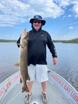 Man standing on a boat holding a large fish with one hand; lake and sky in the background.