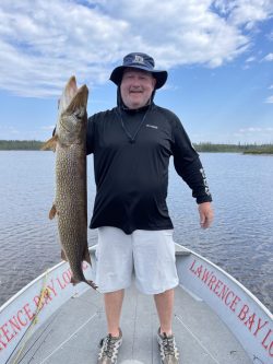 Man standing on a boat holding a large fish upright with one hand, smiling at the camera on a lake.