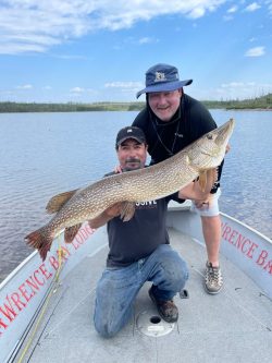 Two men on a boat hold a large fish together on a lake under a blue sky, with lodge branding on the boat's edge.