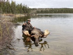 A person in camouflage poses with a rifle beside a large moose in shallow lake water, with a forest in the background.