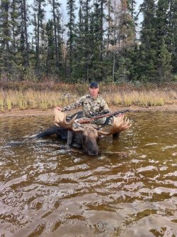 A person in camouflage holds a rifle, posing with a large moose in shallow water near a forested area.