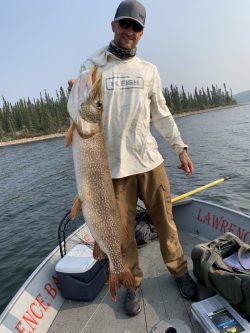 Man standing on a boat holding a large fish, with water and trees in the background.