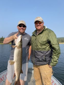 Two men on a boat smiling, one holding a large fish by the mouth; lake and trees in the background.