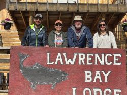 Four people stand behind a large red sign that reads "Lawrence Bay Lodge" with a fish illustration.