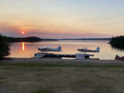 Two floatplanes are docked at a calm lakeshore during sunset, with trees and distant hills in the background.